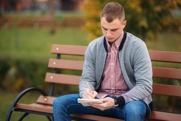 A cute student sits on the bench writes down his thoughts in his notebool using a pensil