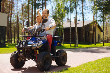 Crazy driving. Delighted young father smiling and driving an all-terrain vehicle with his son