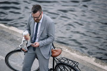 businessman with coffee to go and smartphone standing with bike on quay