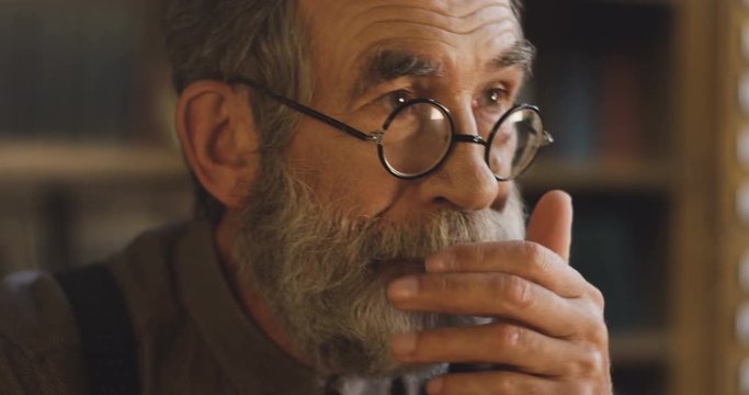 Close up of the old man with beard, gray hair and in glasses thinking and worrying in the library. Portrait shot.