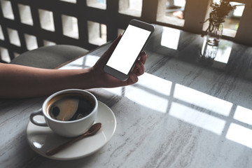 Mockup image of woman's hand holding white mobile phone with blank desktop screen and coffee cup on table in cafe