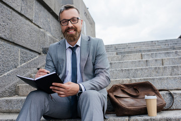 businessman with leather bag and coffee to go writing in diary and sitting on stairs