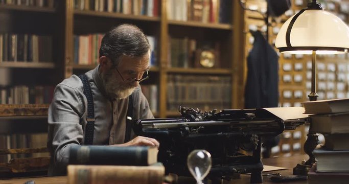 Caucasian old male professor in glasses typing on the typewriter while sitting in the dark ancient library room with a lantern.