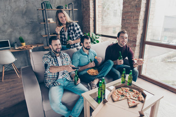 Four attractive, stylish guys with modern hairstyle in jeans watching television, using remote controller, drinking lager eating snacks, sitting in living room, close friends having fun together