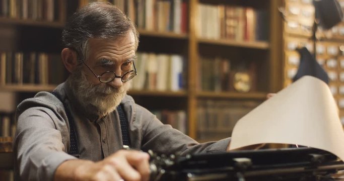 Old Caucasian man in glasses typing on the typewriter and reading what he wrote after that while sitting in the dark ancient library cabinet.