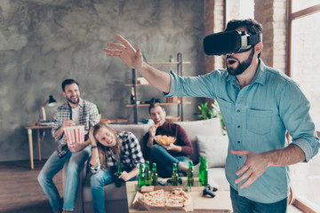 Portrait of amazed attractive bearded guy in vr-headset watching video trying to touch something with arms, his happy friends eating chips, popcorn on blurred background laughing at him
