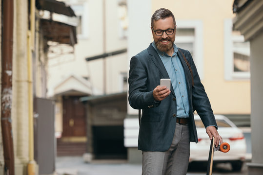 stylish bearded man with longboard using smartphone in city