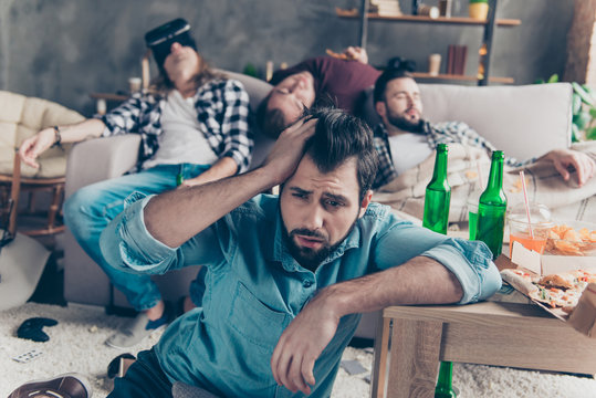 What we did last night? Bearded, upset, unhappy guy suffering from headache after night events, woke up on the floor holding hand on head, his drunk friends sleeping on couch on blurred background