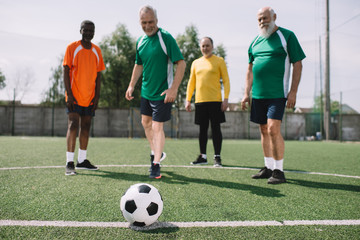 selective focus of football ball and multicultural elderly men on green football field
