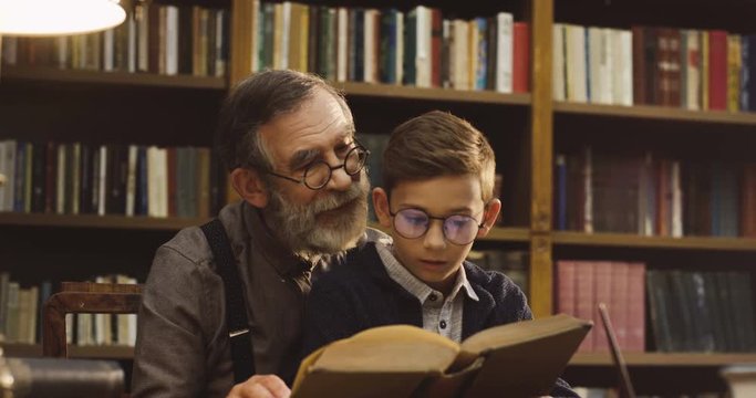 Old grandfther and his teen grandson in glasses sitting in the library and man reading a book for a boy.