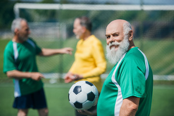 selective focus of old bearded man with football ball and friends behind on field