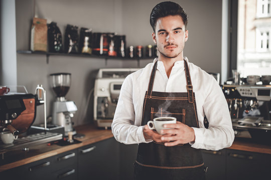 Professional barista in apron holding cup of coffee at cafe