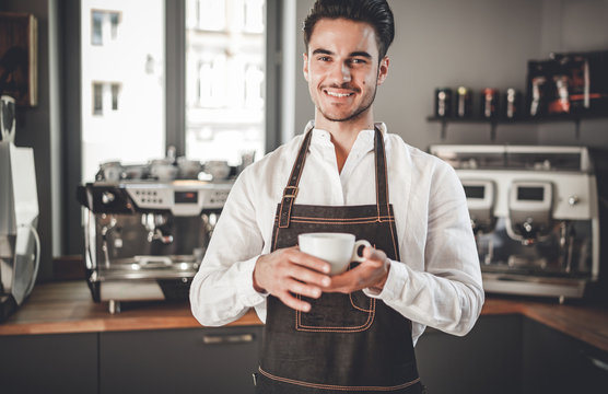 Professional barista in apron holding cup of coffee at cafe