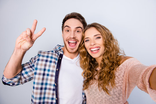 Self portrait of peaceful friendly couple shooting selfie on front camera gesturing v-sign keeping mouth open having online meeting isolated on grey background