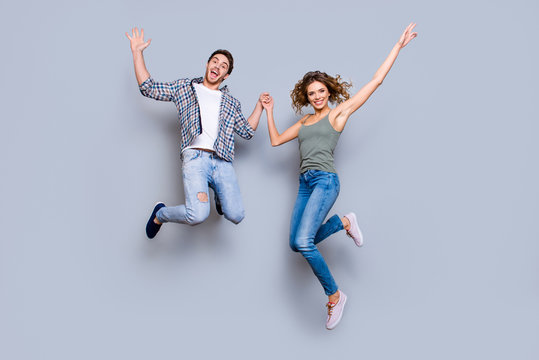 Portrait of funky foolish couple in jeans sneakers jumping in air holding hands up enjoying time together isolated on grey background. Move motion concept