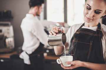 Barista woman pouring milk to coffee cup standing at the bar in cafe