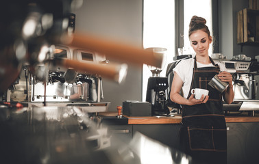 Barista woman pouring milk to coffee cup standing at the bar in cafe