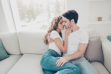 Portrait of sexy romantic couple in casual denim clothes cuddling sitting on sofa indoor kissing with close eyes. Touch gentle satisfaction love story concept