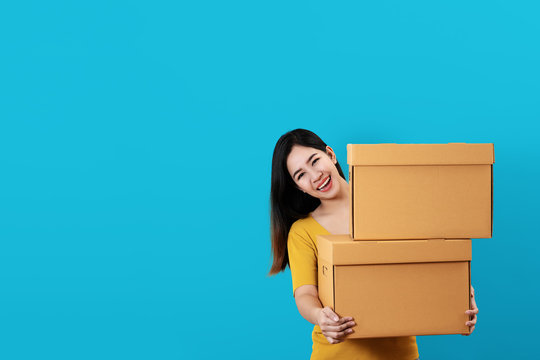 Portrait of young asian entrepreneur happy smiling and holding or carrying pile of boxes in hands with blue isolated background and copy space. Asian woman move to new home or ecommerce owner concept.