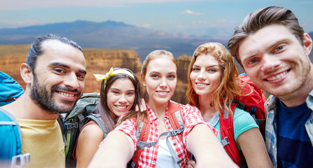 Obraz premium technology, travel, tourism, hike and people concept - group of smiling friends walking with backpacks taking selfie by smartphone or camera in woods