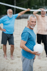 selective focus of smiling elderly man with volleyball ball looking at camera with old friends...