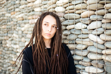 Portrait of a beautiful lonely girl with dreadlocks looking away, against a wall of gray stones. A...