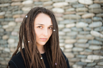Portrait of a beautiful lonely girl with dreadlocks looking at the camera, against a wall of gray stones. Women hipster in black clothes.