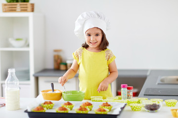 family, cooking, baking and people concept - happy little girl in chefs toque making batter for muffins or cupcakes at home kitchen