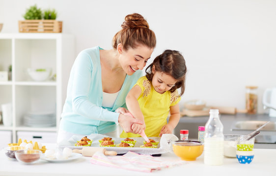 Family, Cooking And People Concept - Mother And Little Daughter Making And Decorating Cupcakes With Cream Frosting At Home Kitchen