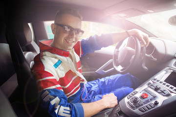 A young handsome muscular male racer in a commenced ride and posing on a car in summer sunny weather