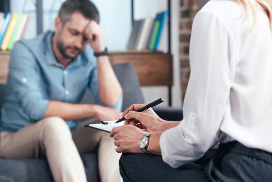 cropped image of female counselor writing in clipboard while depressed male patient sitting on sofa at office