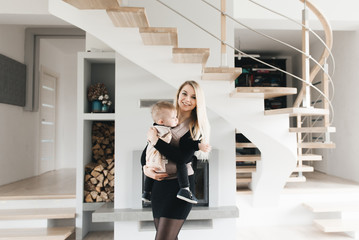 Stylish young mom keeps in ergo backpack of her cute year-old baby at home in a beautiful interior Convenience for mom.