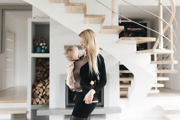 Stylish young mom keeps in ergo backpack of her cute year-old baby at home in a beautiful interior...