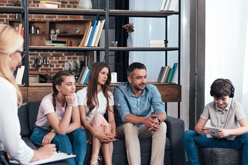 smiling boy in headphones using smartphone while family looking at him on therapy session by female counselor in office