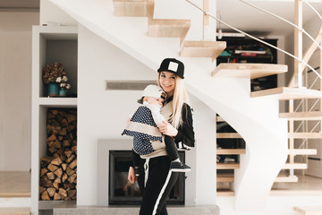 Stylish young mom keeps in ergo backpack of her cute year-old baby at home in a beautiful interior Convenience for mom.