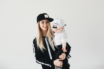 Stylish young mom in cap holding her cute one year old baby isolated on white background.