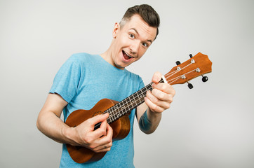 A young smiling guy plays on a ukulele on a light background.