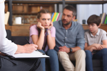 cropped image of female counselor writing in clipboard while family sitting on sofa on therapy session