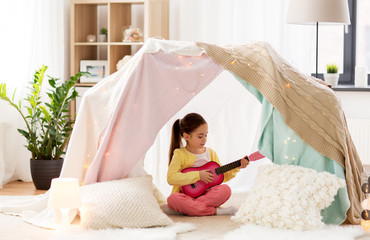 Obraz na płótnie Canvas childhood and hygge concept - happy little girl playing toy guitar in kids tent at home