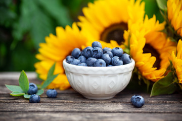 Ripe blueberries in white bowl with sunflower bouquet on wooden table, summer theme