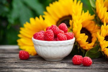 Ripe raspberries in white bowl with sunflower bouquet on wooden table, summer theme