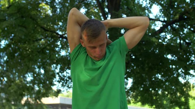 Handsome runner warming up before jogging in summer park. Healthy active man stretching arms and neck before running and training outdoors. Close-up. Slow motion.