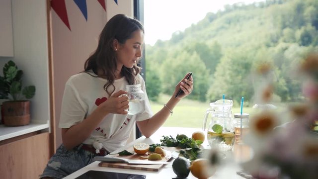 Attractive young woman in a stylish white t-shirt drinks lemonade from a mason jar and browses social networks via her phone while sitting in a cozy modern kitchen. Modern communication, free time.