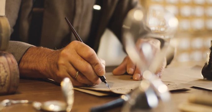 Close up of the old male hands writing a letter with ink on the wooden table in the ancient style.
