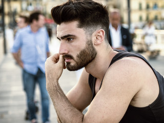 Fototapeta na wymiar Attractive sad, worried young man thinking, at sunset on seaside boardwalk or seafront, wearing black tank-top, looking at the sea or ocean