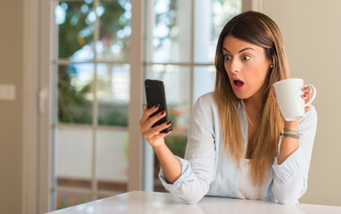 Beautiful young woman looking at the phone suprised and excited while drinking cup of coffee at...