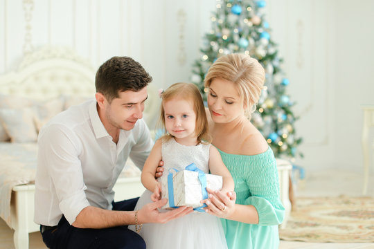 Young mother and father sitting with little daughter keeping present, Christmas tree in background. Concept of winter holidays and hap y famile celebrating New Year.