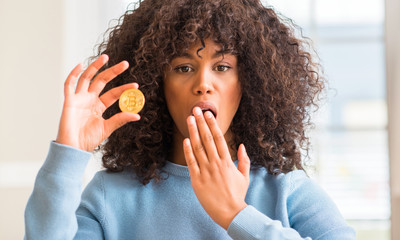 Fototapeta na wymiar African american woman holding golden bitcoin cryptocurrency at home cover mouth with hand shocked with shame for mistake, expression of fear, scared in silence, secret concept
