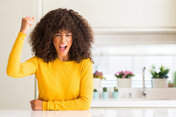 African american woman wearing yellow sweater at kitchen angry and mad raising fist frustrated and furious while shouting with anger. Rage and aggressive concept.