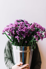 Very nice young woman hiding behind a bucket of beautiful fresh blossoming  purple flowers on the grey background